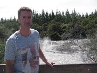 Richark Witbreuk is wearing a pale blue t-shrit with a red pattern. Richard is standing in front of a river with pine trees in the background