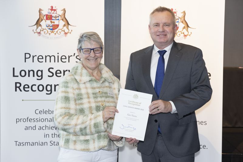 The premier and award recipient pose with certificate in front of banners that read Premier's Long Service Recognition