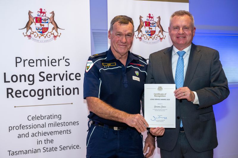 The premier and award recipient pose with certificate in front of banners that read Premier's Long Service Recognition