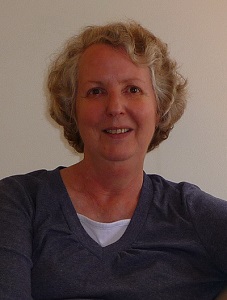 Virginia Case has short, blonde wavy wair and is wearing a steel grey V-neck jumper with a white top