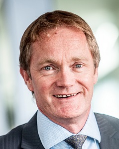 Angus Swindon is smiling. He is wearing a grey suit jacket, collared shirt and grey tie with a light pattern