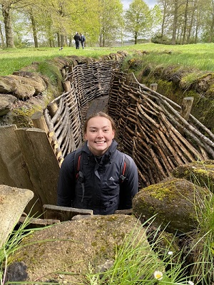 A young woman in a trench that has been reinforced with wicker. The field is green and people are standing in the distance