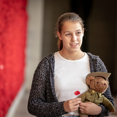 Amelia Hanson standing in front of a memorial wall full of poppies. Amelia is holding a teddy bear in Australian Infantry Force uniform.