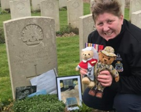 Katie Kelly holding two teddy bears (one dressed as a nurse, the other dressed in military uniform) next to Private John Jack Ernest Johns' headstone